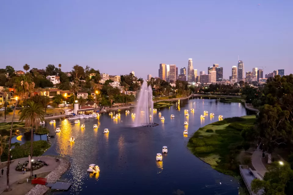 View from above of swan-shaped paddleboats with lights on around artesian fountain in Echo Park Lake with downtown Los Angeles skylien in background at twilight.